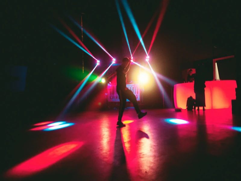 Silhouetted dancer on a dark dance floor surrounded by vibrant red, blue, and green DJ lighting and colorful beams.